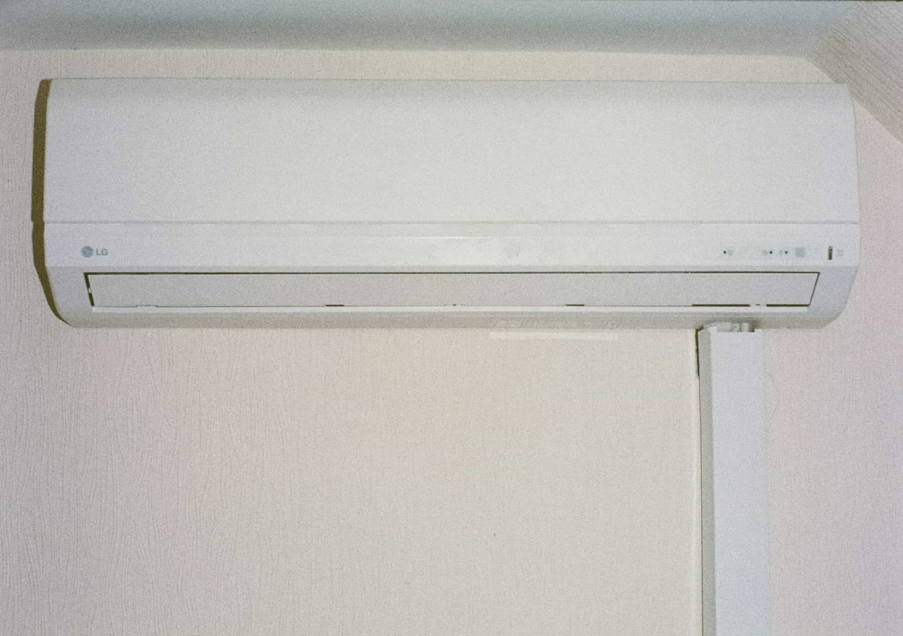 Wall-mounted white air conditioning unit with a vent at the bottom, installed on a light-colored wall.