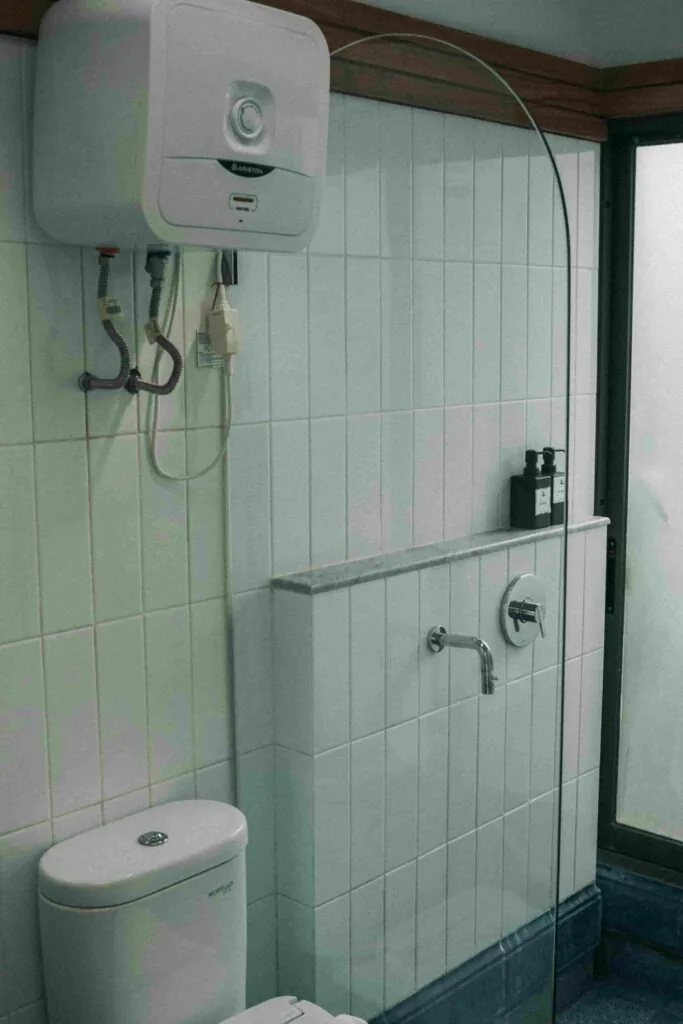 A bathroom with a water heater mounted on the wall above a toilet, a faucet on a tiled partition, and a frosted glass window on the right.