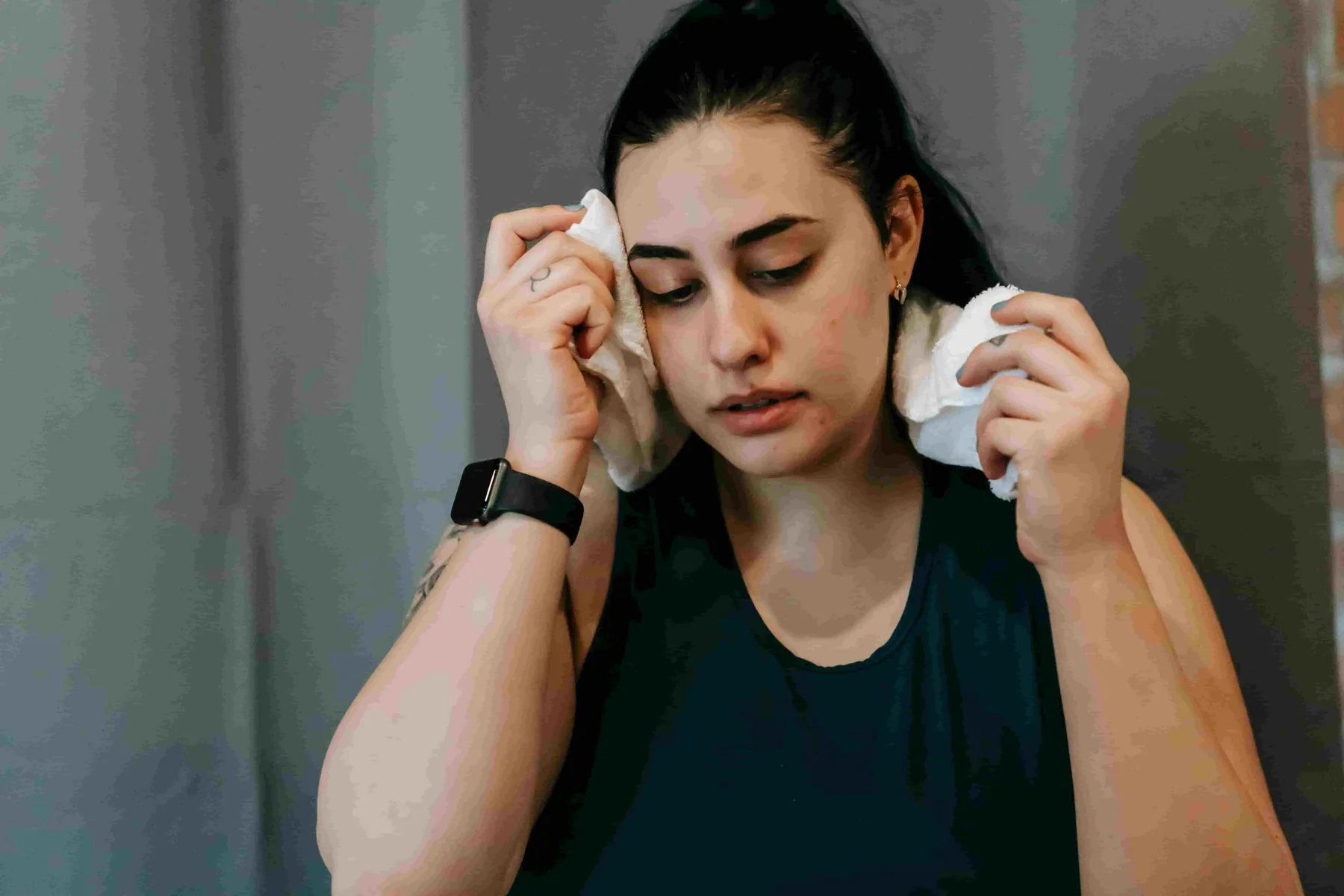 A woman with dark hair holds a towel to each side of her face, wearing a sleeveless top and a smartwatch, standing indoors—perhaps seeking relief while exploring Cooling System Options for Coquitlam Heatwaves.