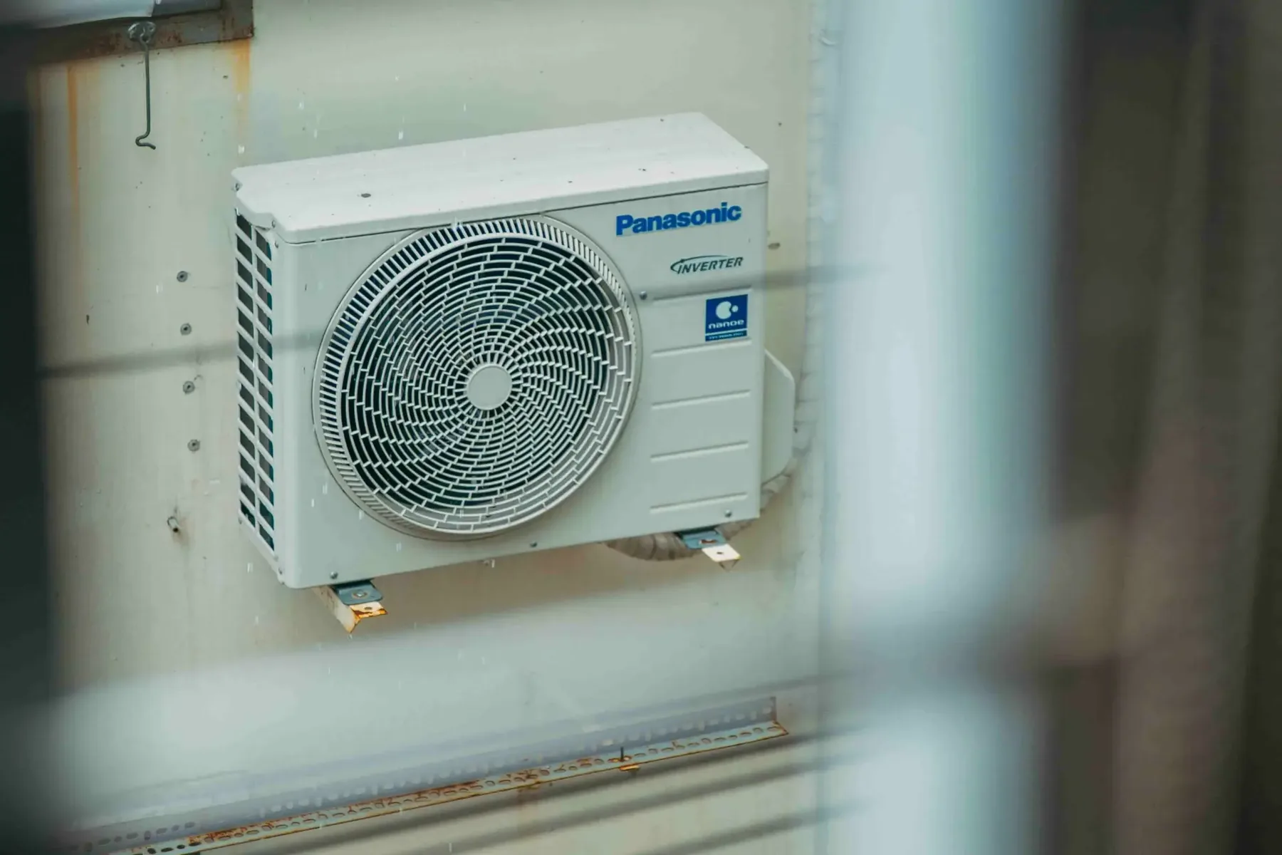 A Panasonic Inverter air conditioning outdoor unit mounted on a wall, with some blurred bars visible in the foreground—a reminder to avoid costly mistakes in HVAC system upgrades.
