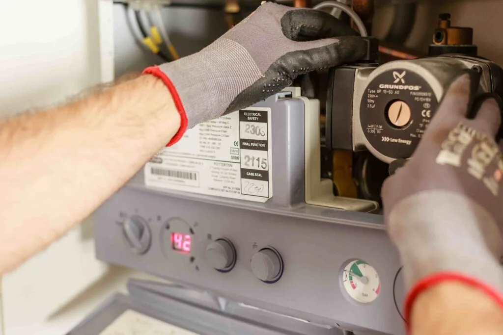 A person wearing gloves adjusts components inside a boiler, with control dials and a digital display visible on the front panel.