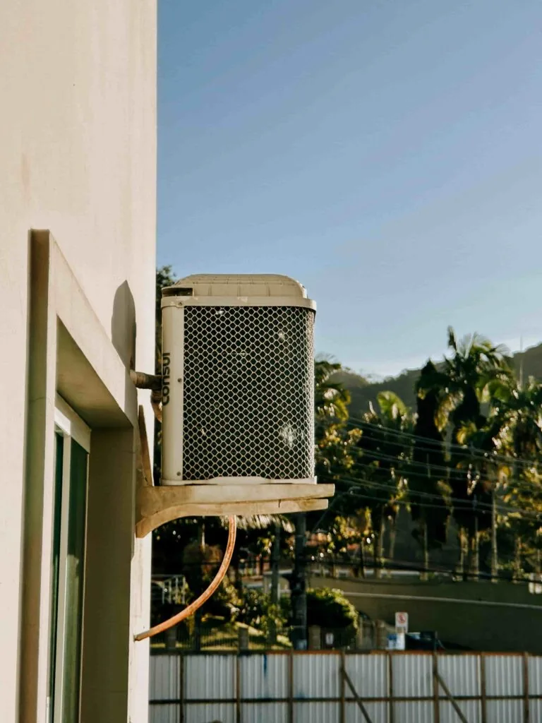 An air conditioning unit is mounted on the exterior wall of a building, with trees, power lines, and a fence visible in the background under a clear sky.