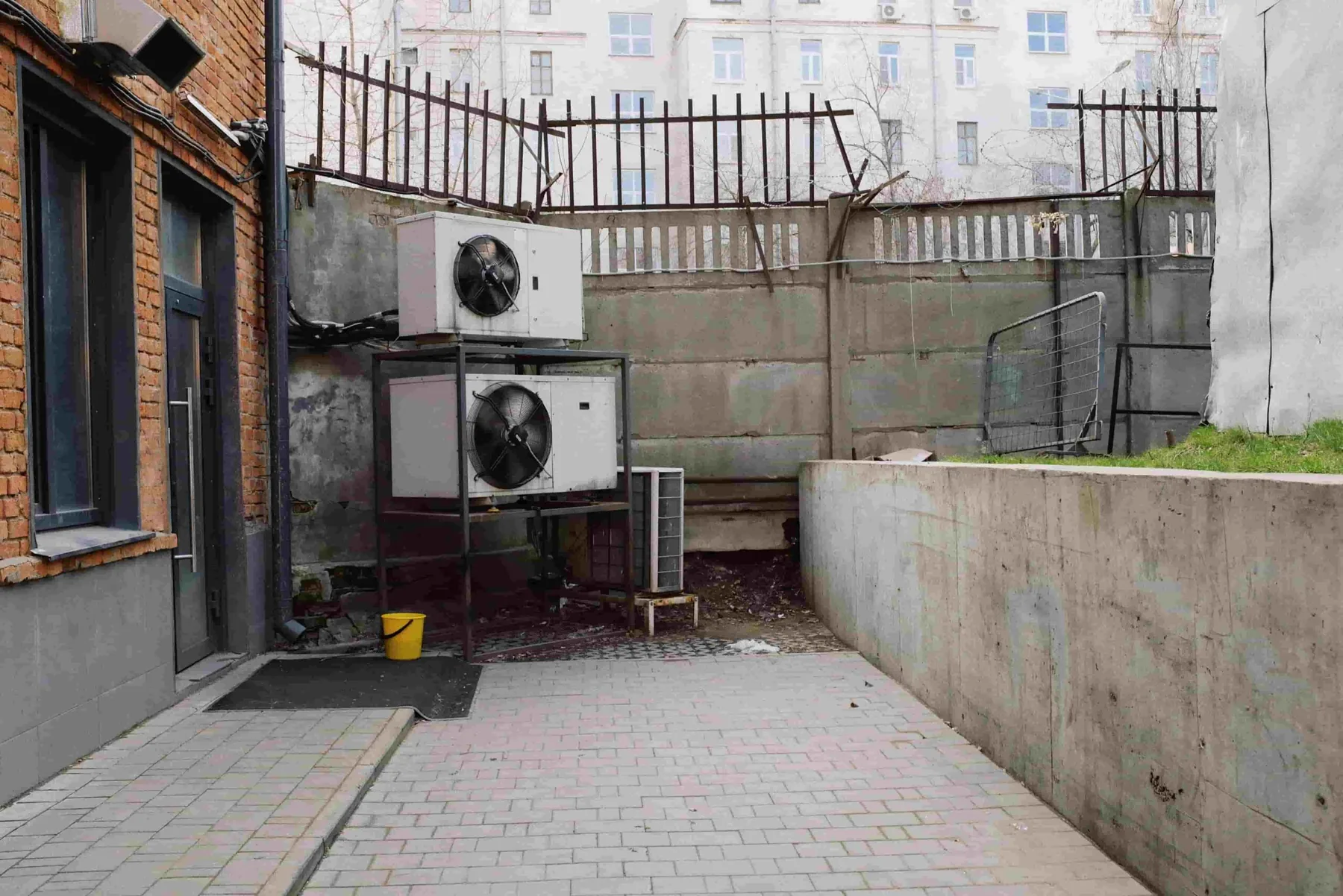 A small paved area with a Heat Pump Setup featuring two large outdoor air conditioning units, a yellow bucket, and a concrete wall topped with metal fencing.