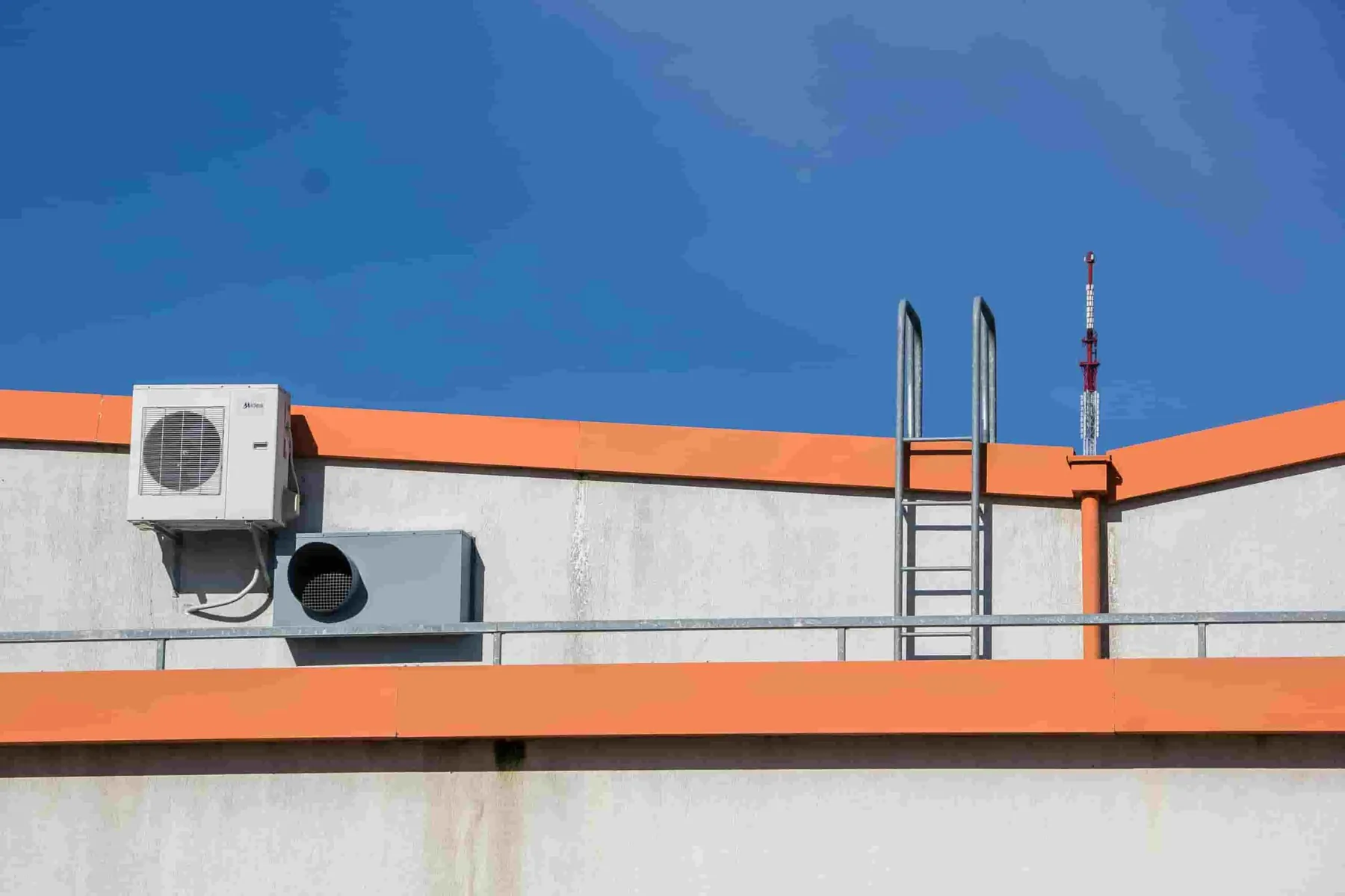 A rooftop with an air conditioning unit, a ventilation duct, a metal ladder, and orange trim under a clear blue sky—an ideal spot for essential HVAC Tune-Ups.