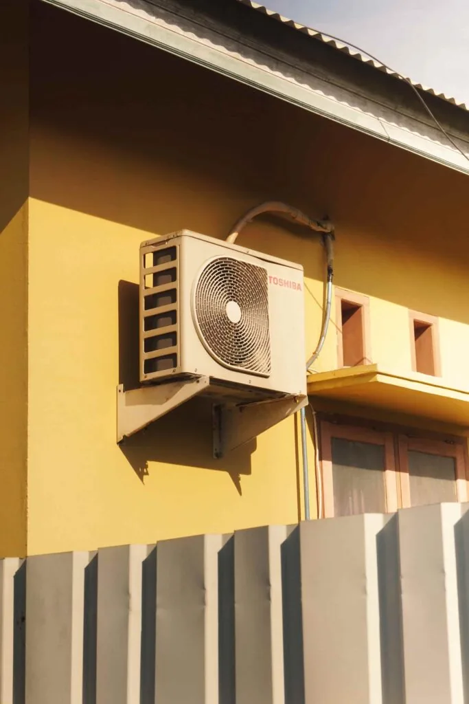An air conditioning unit is mounted on the yellow exterior wall of a building above windows, with a corrugated metal fence in the foreground.