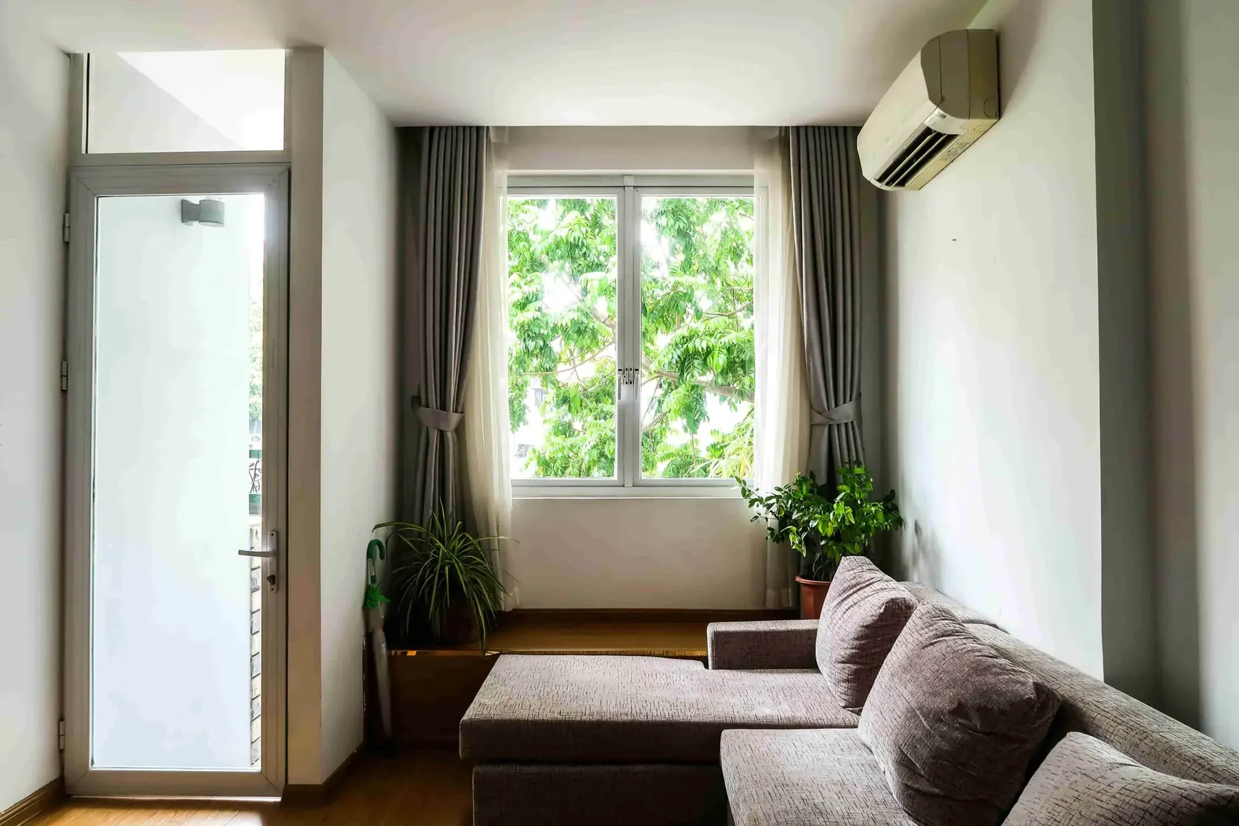A bright living room with a gray sectional sofa, potted plants, a large window with curtains, an air conditioner—perfect for those with questions for HVAC contractors in Vancouver—and a glass door.