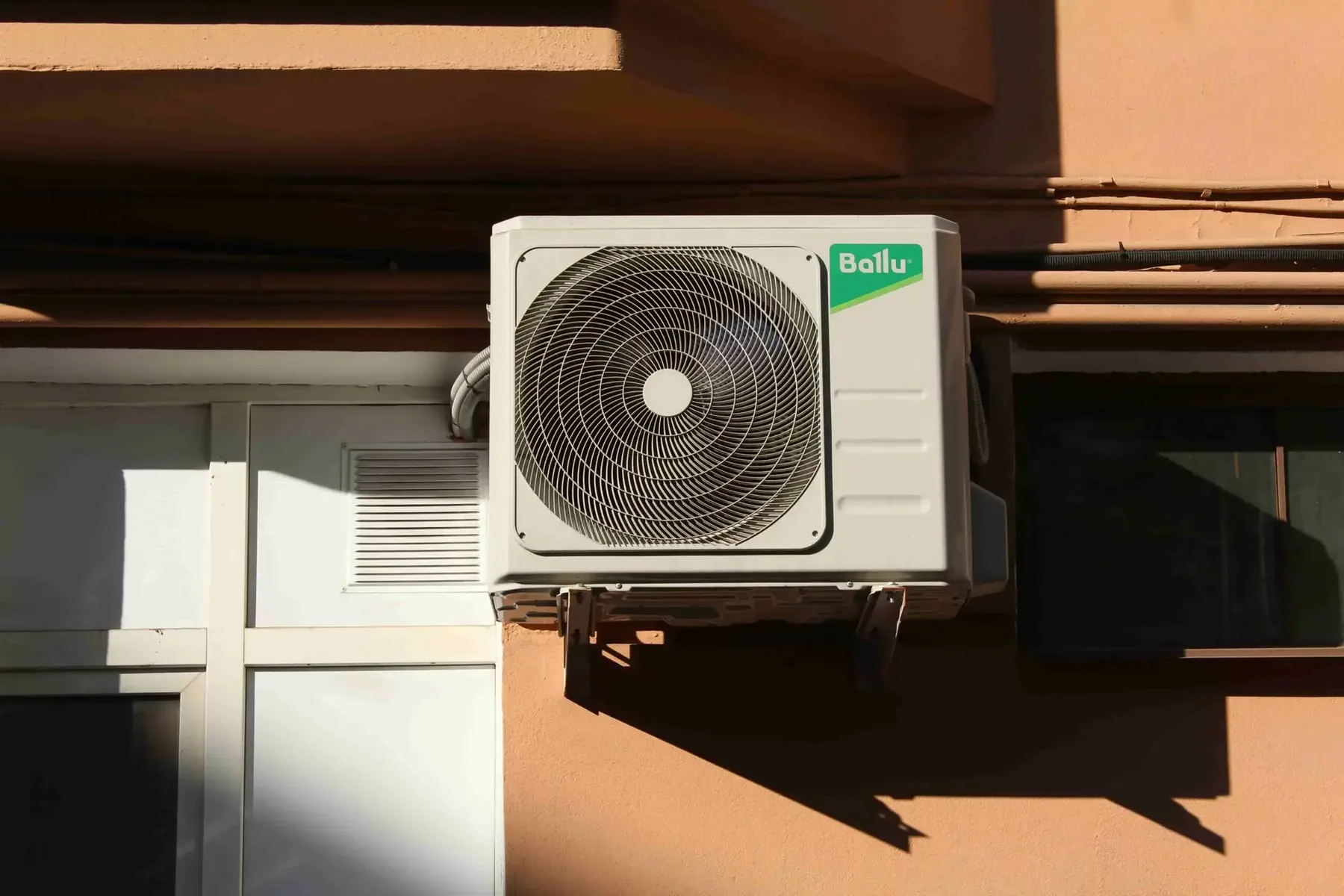 Outdoor air conditioning unit mounted on a peach-colored wall beside a window, featuring visible electrical wiring and a Ballu brand label—ideal for those wondering, "Where Can I Get Ductless Mini-Split Installation in Vancouver?.