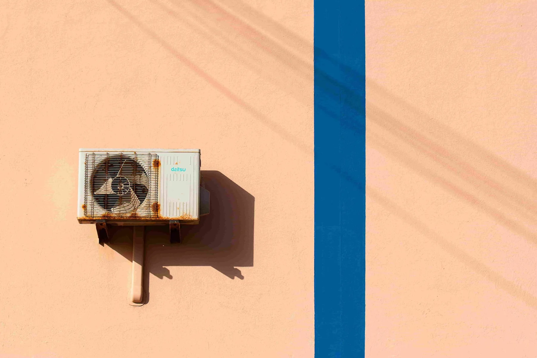 A rusted air conditioning unit mounted on a peach-colored wall with a vertical blue stripe and shadow lines hints at the importance of regular maintenance, like seeking where to get seasonal HVAC inspections in New Westminster.