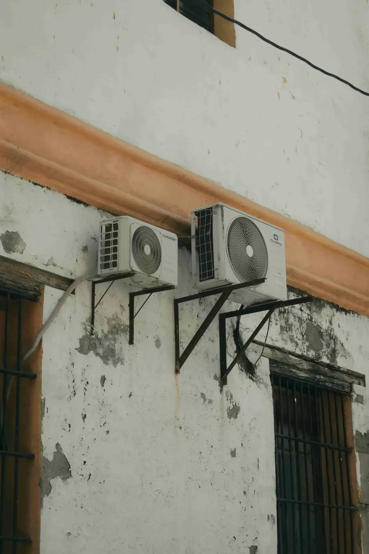 Two outdoor air conditioning units mounted on a weathered, peeling white wall between barred windows—wondering Who Offers Ductless Mini-Split Installation in Langley BC? This setup shows what's possible for your space.